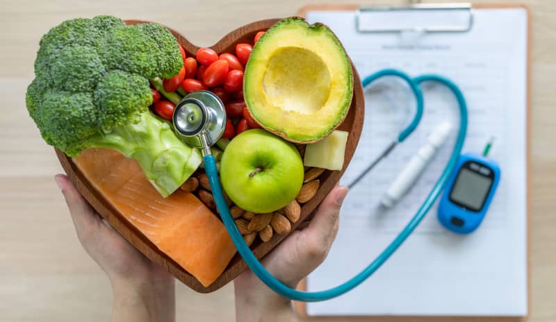 Plato en forma de corazón con alimentos saludables como brócoli, salmón, aguacate, manzana, frutos secos y tomates; junto a un estetoscopio y un glucómetro sobre una hoja clínica, simbolizando el control del colesterol elevado mediante una dieta equilibrada.