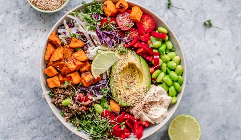 Bowl con aguacate, boniato, edamame, hummus, quinoa, verduras frescas y semillas, ejemplo colorido de desayuno saludable.