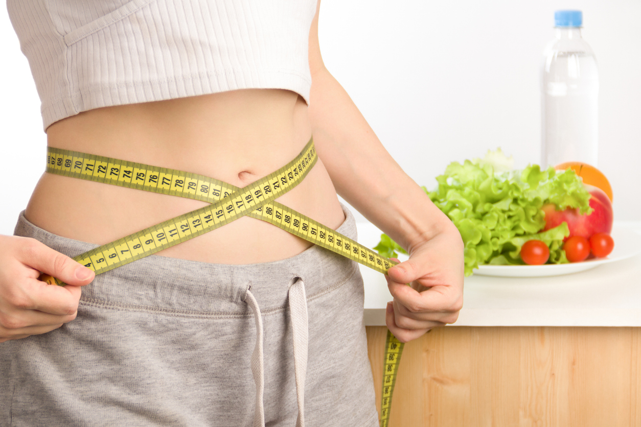 Mujer con un top corto que deja visible su ombligo, midiendo su cintura con cinta métrica amarilla, imagen asociada al objetivo de bajar 4 kilos en un mes.