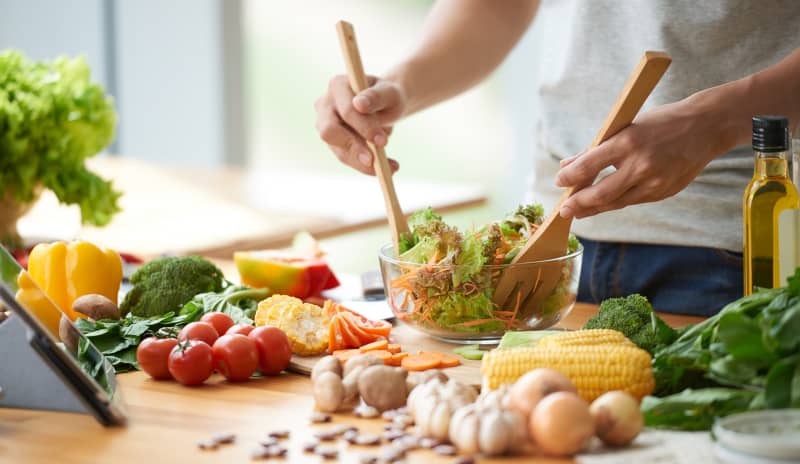 Un hombre prepara una ensalada con verduras frescas en su cocina en la que se ven ingredientes como ajos, frutos secos, pimientos, tomates o aceite y una tablet que parece estar viendo mientras cocina, ejemplo de alimentación saludable en distintos tipos de dietas.