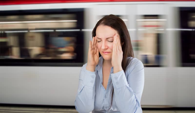 Mujer con expresión de malestar y ansiedad, sentada frente a un tren en movimiento, llevándose las manos a la cabeza, representando síntomas comunes en personas con agorafobia en espacios públicos o transitados.