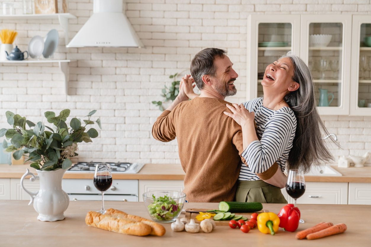 Pareja adulta sonriendo mientras baila en la cocina de casa mientras cocina y se ven verduras y hortalizas coloridas sobre la mesa, ejemplo de convivencia emocional en el modelo de relación living apart together.