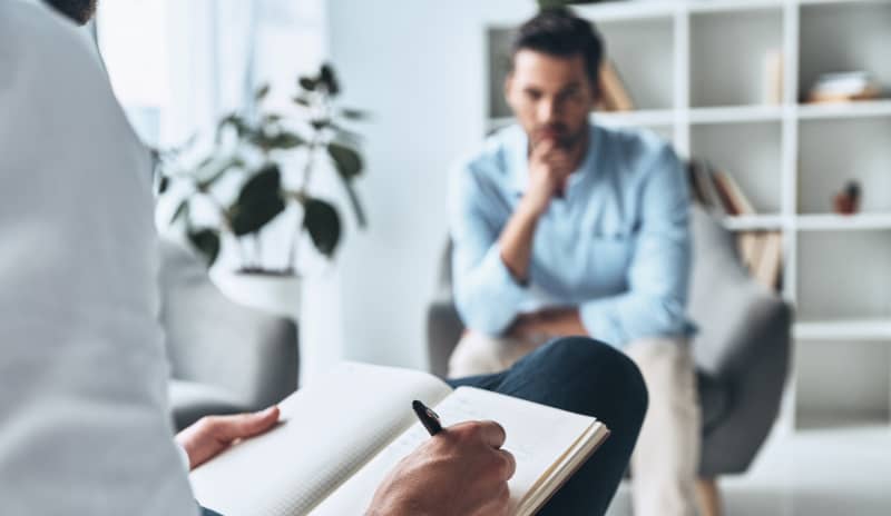 Hombre sentado en consulta psicológica mientras el profesional toma notas en una libreta, en una sesión relacionada con la mitomanía