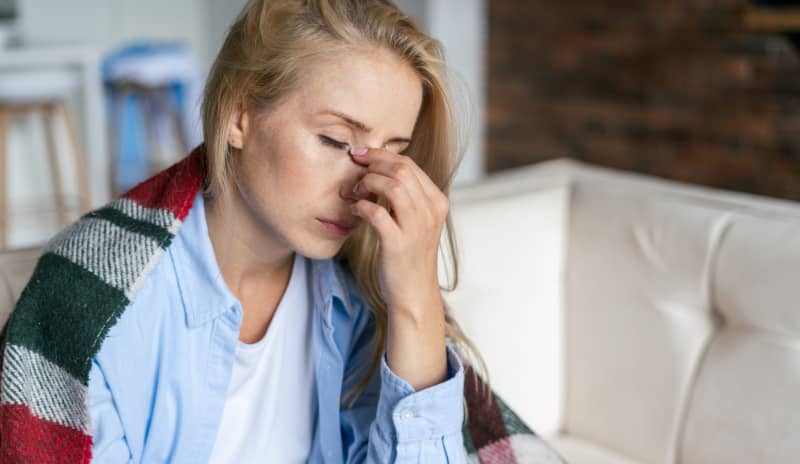 Mujer sentada en el sofá con una manta por encima presionando el puente de la nariz con expresión de molestia, mostrando síntomas compatibles con rinitis no alérgica como congestión y presión facial