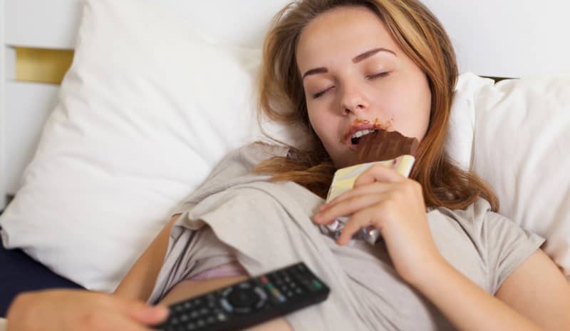 Mujer dormida comiendo una tableta de chocolate en una superfecie cómoda y con el mando de la tele en una mano, conducta propia de las personas que padecen sonambulismo.