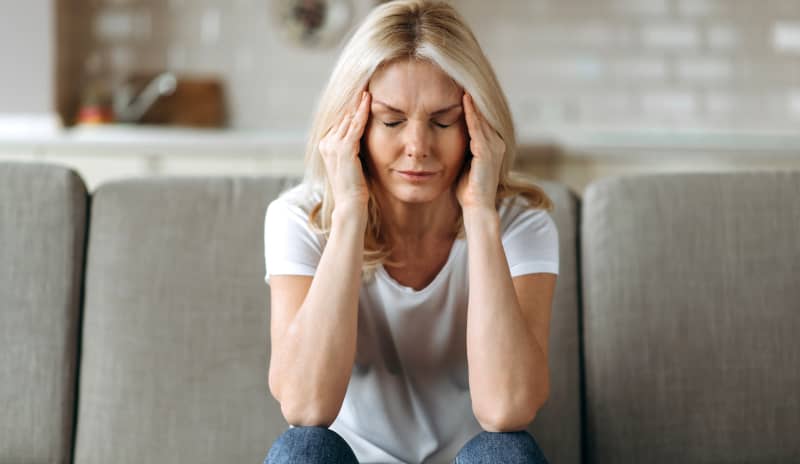 Mujer sentada en el sofá de una casa con los ojos cerrados y las manos en las sienes, gesto común ante el dolor de cabeza por cefalea tensional crónica.