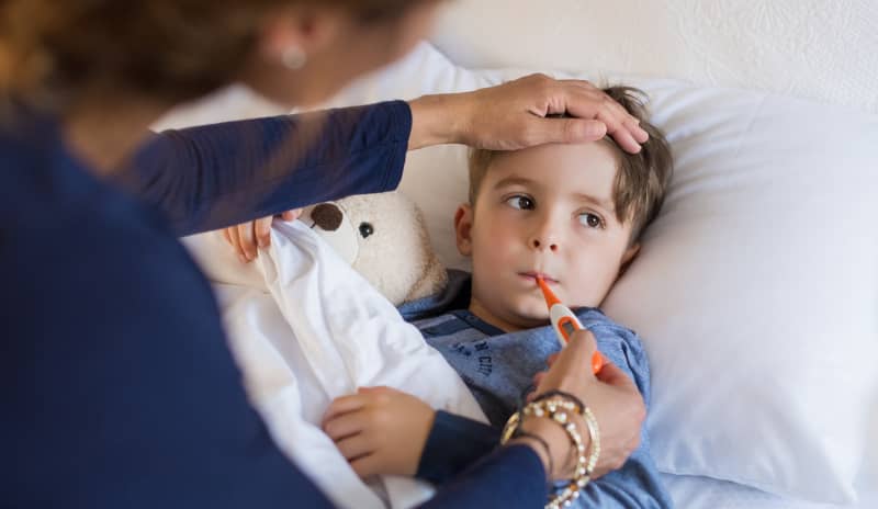 Niño en la cama con un termómetro digital en la boca mientras un adulto le toca la frente, escena relacionada con la medición de la temperatura corporal.