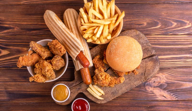 Mesa con comida rápida: pollo frito, patatas fritas, perritos calientes y una hamburguesa, representando varios de los 10 peores alimentos para el corazón por su alto contenido en grasas saturadas y sodio