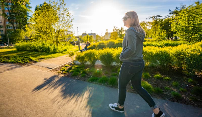 Mujer caminando por un parque urbano en un día soleado, rodeada de vegetación, ilustrando los beneficios de caminar para la salud, la actividad física diaria y el bienestar mental