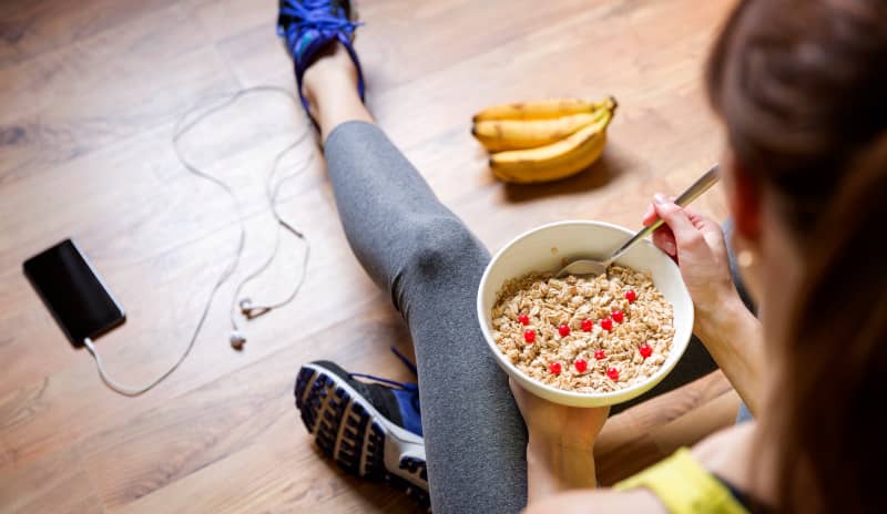 Una chica sentada en el suelo y vestida de deporte sostiene un tazón de avena con leche, en lo que podría ser un desayuno para ganar masa muscular, mientras a su derecha se ve un racimo de plátanos y a su izquierda se ve un móvil con un cable que parecen ser los cascos para escuchar música. La fotografía está tomada en un plano picado.