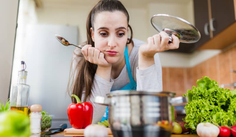Mujer en la cocina con gesto de indecisión frente a una olla y verduras frescas, imagen asociada a la dieta Dukan y a la planificación de comidas altas en proteínas
