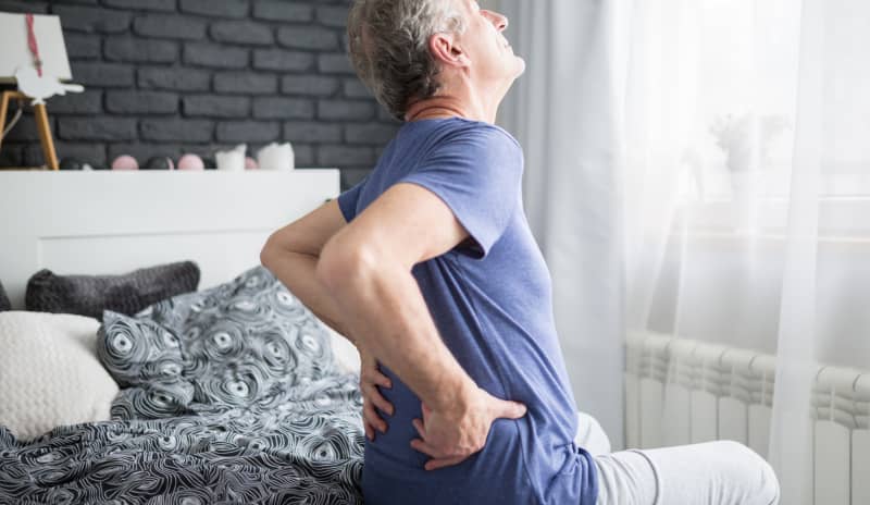 Hombre sentado al borde de la cama sujetándose la parte baja de la espalda con gesto de molestia, mostrando síntomas claros de dolor lumbar
