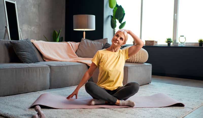 Una mujer sentada en una esterilla en el salón de una casa y vestida de deporte con las piernas cruzada, realiza ejercicios de estiramiento cervical para evitar los mareos cervicales.