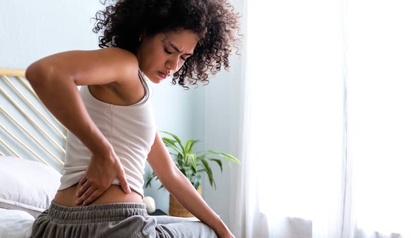 Mujer de tez oscura y pelo muy rizado sentada al borde de la cama presionándose la zona lumbar con gesto de molestia, ilustrando síntomas de dolor de espalda baja