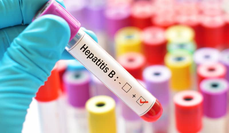 Mano con guante sostiene un tubo de sangre con etiqueta “Hepatitis B” y resultado positivo, con varios tubos de muestras de colores desenfocados al fondo