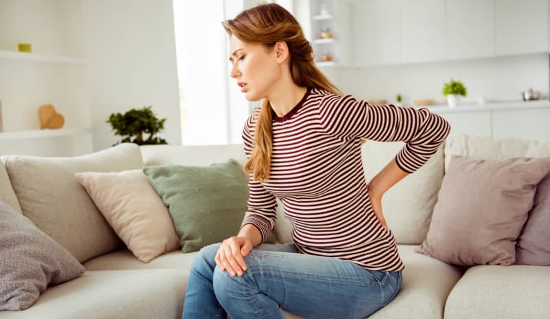 Mujer sentada en el sofá con gesto de dolor y la mano en la zona lumbar, representando uno de los síntomas de cálculos renales