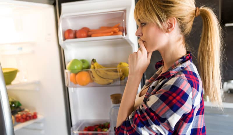 Mujer pensativa frente al refrigerador abierto con frutas y verduras, evaluando opciones de alimentación saludable para perder peso