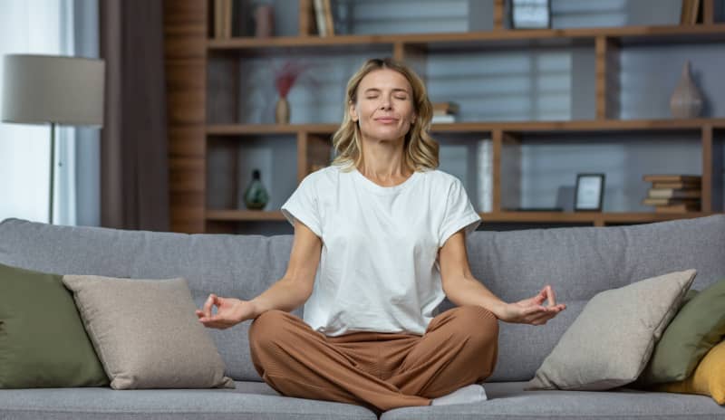 Mujer practicando meditación en posición de loto en el sofá de su casa con los ojos cerrados, una técnica de autocuidado efectiva para superar el burnout parental.