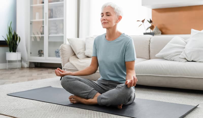 Mujer madura practicando meditación en casa sobre una esterilla de yoga, con expresión serena y los ojos cerrados