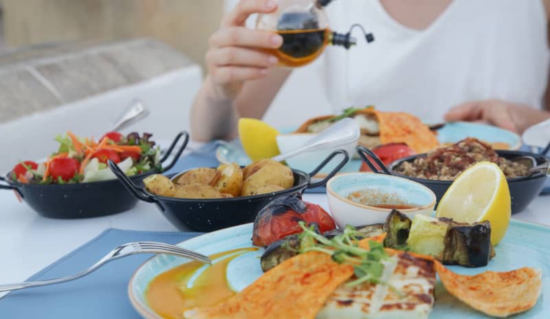 Persona en una mesa con platos de verduras asadas, ensalada, patatas y limón aliñando con aceite de oliva , ejemplo de comida mediterránea rica en alimentos antiinflamatorios.