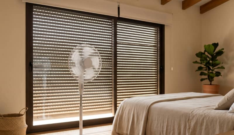 Ventilador de pie frente a una cama y una ventana con la persiana entreabierta en un dormitorio, una opción habitual cuando hay que gestionar cómo dormir con calor