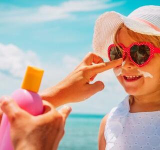 Mano de alguien aplicando protector solar en la cara de una niña con sombrero y gafas de sol en la playa, imagen relacionada con el uso de protector solar para niños