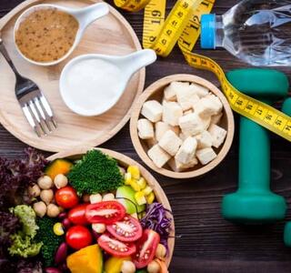Platos de comida saludable como una ensalada, un bol de tofu y especias, junto a un par de pesas, una botella de agua y una cinta métrica que envuelve todos los objetos, elementos relacionados con el objetivo de bajar 4 kilos en un mes.