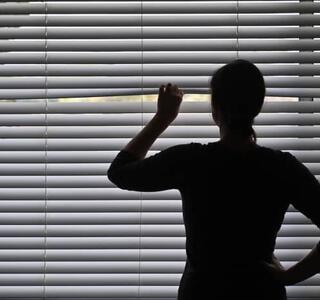 Persona en silueta observando hacia el exterior a través de una persiana cerrada, simbolizando el aislamiento y el temor a salir de casa característico de la agorafobia.