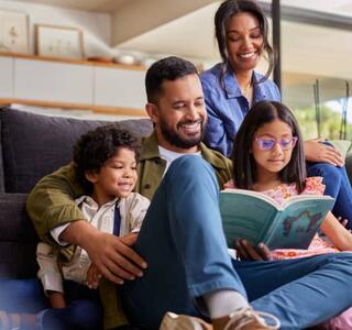 Familia sonriente reunida en casa, con el padre leyendo un libro a sus dos hijos mientras la madre los observa sentada en el sofá, en un ambiente cálido y relajado, representando el día de la familia.