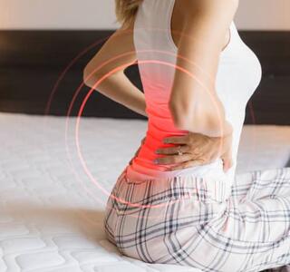 Mujer sentada en la cama sujetándose la parte baja de la espalda con una zona roja destacada, representando un episodio de dolor lumbar