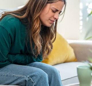 Mujer joven sentada en el sofá de una casa sujetándose el abdomen con gesto dolor, en representación del dolor de vientre bajo sin menstruación