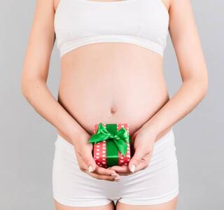 Mujer embarazada con ropa interior blanca sosteniendo un pequeño regalo sobre el vientre, imagen relacionada con el embarazo críptico
