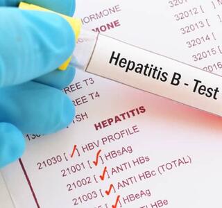 Guante médico sostiene un tubo de sangre con etiqueta “Hepatitis B - Test” sobre un informe de laboratorio con resultados de hepatitis B