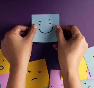 Manos colocando una nota adhesiva con una cara sonriente entre otras notas con expresiones emocionales variadas, imagen que representa la salud mental, el reconocimiento de emociones y el bienestar emocional.