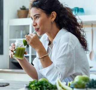 Mujer bebiendo un batido verde en la cocina, rodeada de frutas y verduras frescas, como parte de hábitos saludables para perder peso