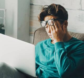 Joven con gafas sentando en un sillón de una casa frente al ordenador portátil con gesto de agotamiento y preocupación, representando la ansiedad anticipatoria