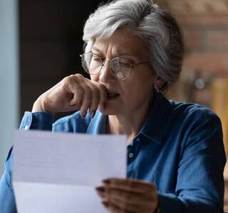 Mujer mayor con gafas leyendo un documento con gesto de preocupación en su cocina, reflejando la ansiedad financiera que generan las facturas y notificaciones económicas