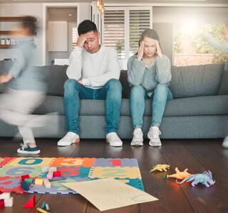 Pareja de padres exhaustos sentados en el sofá con gesto de agotamiento mientras sus hijos corren a su alrededor, ilustrando el burnout parental