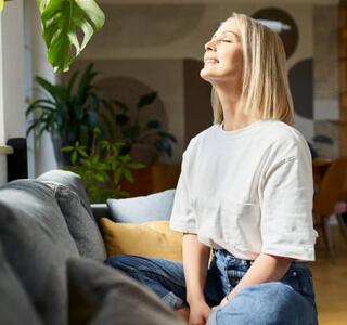 Mujer relajada sentada en el sofá con los ojos cerrados y expresión serena en un salón luminoso con plantas, representando la calma y el bienestar del dopamine detox