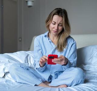 Mujer joven sentada en la cama mirando su móvil con expresión de preocupación y ansiedad, representando la nomofobia o dependencia del teléfono