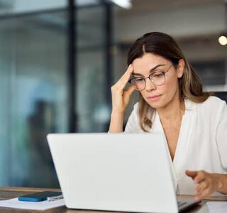 Mujer profesional con gafas frente al ordenador portátil en una oficina con expresión de duda, representando el síndrome del impostor en el trabajo