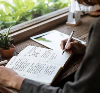 Manos y brazo de espaldas de una mujer escribiendo a mano en un diario emocional, en representación del journaling terapéutico, en una mesa de madera junto a una ventana con plantas y luz natural.