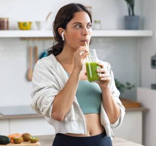 Mujer apoyada en la encimera de una cocina bebiendo un batido verde de frutas y verduras vestida de deporte, un ejemplo de microhábitos de alimentación saludable