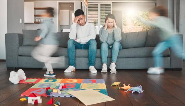 Pareja de padres exhaustos sentados en el sofá con gesto de agotamiento mientras sus hijos corren a su alrededor, ilustrando el burnout parental
