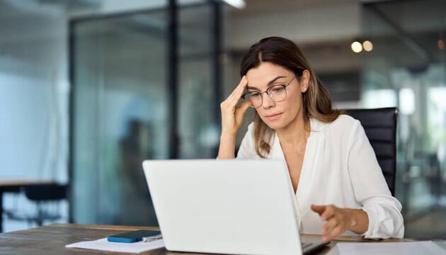 Mujer profesional con gafas frente al ordenador portátil en una oficina con expresión de duda, representando el síndrome del impostor en el trabajo