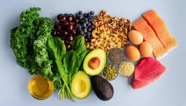 Vista cenital de alimentos antiinflamatorios como salmón, atún, kale, espinacas, aguacate, arándanos, nueces, semillas, huevos y aceite de oliva sobre fondo claro