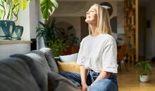 Mujer relajada sentada en el sofá con los ojos cerrados y expresión serena en un salón luminoso con plantas, representando la calma y el bienestar del dopamine detox