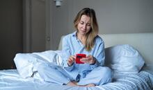 Mujer joven sentada en la cama mirando su móvil con expresión de preocupación y ansiedad, representando la nomofobia o dependencia del teléfono