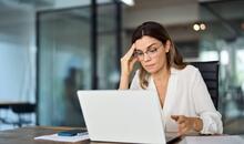 Mujer profesional con gafas frente al ordenador portátil en una oficina con expresión de duda, representando el síndrome del impostor en el trabajo