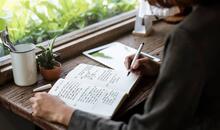 Manos y brazo de espaldas de una mujer escribiendo a mano en un diario emocional, en representación del journaling terapéutico, en una mesa de madera junto a una ventana con plantas y luz natural.